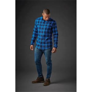 A man wearing a blue and black checkered button-up shirt, paired with jeans and brown shoes, standing against a dark background.