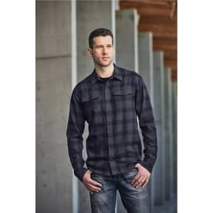 A man wearing a black and grey checked button-up shirt, standing against a concrete wall. The shirt has a logo.