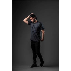 A model wearing a short-sleeve, dark grey T-shirt paired with black pants against a dark background. The shirt features a logo.