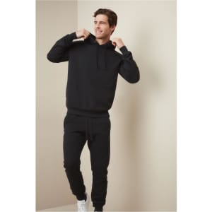 A black unisex sweatshirt with a hood, worn by a smiling man adjusting the hood, featuring a logo.