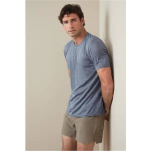 A man is wearing a blue recycled sports T-shirt, standing against a light wall, paired with beige shorts.