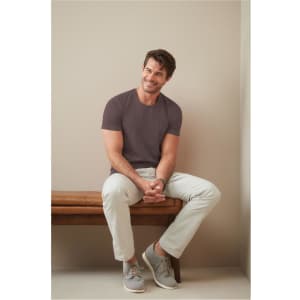 A model sits on a bench, wearing a mauve crew neck t-shirt and light-coloured trousers with grey sneakers.