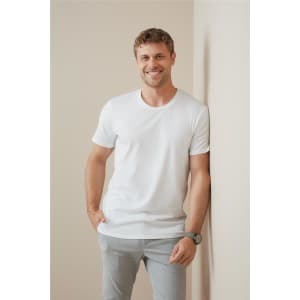 A man in a white crew neck t-shirt leans against a wall, smiling and wearing light grey pants.