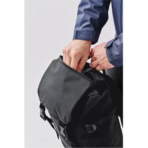 A black backpack with a zippered opening, being handled by a person in a blue shirt. It features adjustable straps and buckles.
