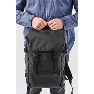 A black backpack with adjustable straps, held by a person wearing a blue jacket. It features a front pocket and a logo.