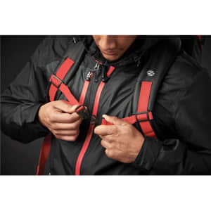 A black backpack with red straps is being fastened by a person in a dark setting. The backpack has a logo on it.