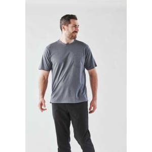A man stands wearing a grey short sleeve t-shirt with a pocket, paired with dark pants. The shirt features a logo.