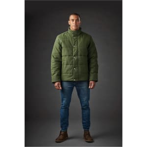 A man stands wearing a dark green thermal jacket with a button-up front, paired with jeans and boots.