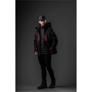 A man in a black jacket with red accents layered over a quilted jacket, wearing a cap and dark pants.