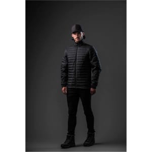 A man wearing a black puffer jacket stands against a dark background, complemented by black trousers and boots.