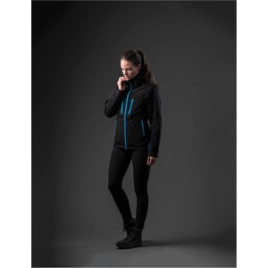 A woman wearing a black jacket with blue accents, styled with black leggings and boots, stands against a dark background.