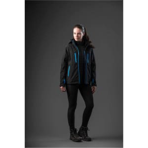 A woman in a black jacket with blue accents stands against a dark background, wearing black leggings and boots.