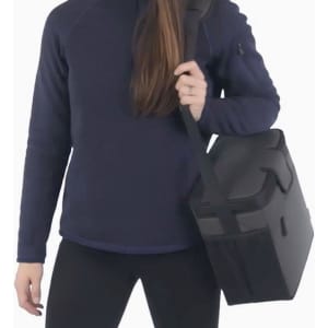 A cooler bag with a grey and black design being held by a person wearing a navy sweater. The bag has a strap and a handle.