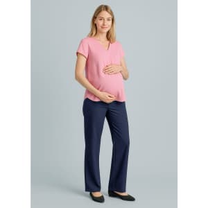 A pregnant woman wears a pink top and navy maternity pants, standing with one hand on her belly. The background is neutral.