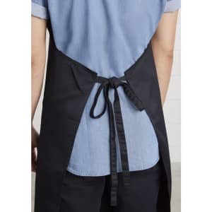 A bib apron in black, featuring a tie at the back and worn over a light blue shirt.