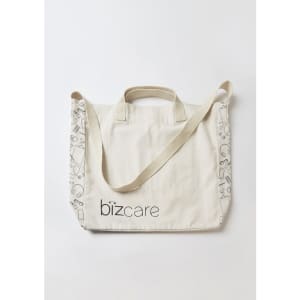 A cream tote bag with a strap, featuring a logo and patterned detailing along the edges.