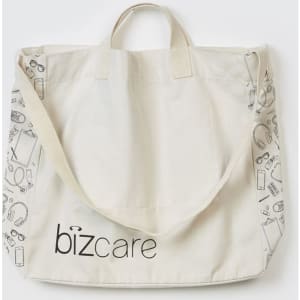 A beige unisex tote bag featuring a repeat pattern of various items and a logo. It has short handles and a long strap.
