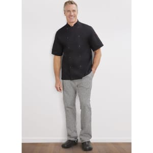 A man stands smiling in a black short-sleeve chef jacket and striped grey pants, with black shoes and a logo on the jacket.