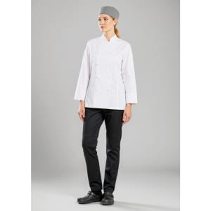 A woman in a white long sleeve chef jacket, black pants, and a grey chef's hat, standing against a grey background.