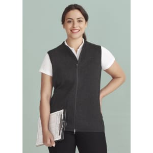 A woman wearing a black zip-front vest over a white shirt, holding a clipboard, against a light green background.