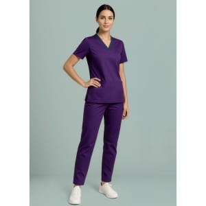 A woman wearing a purple scrub top and matching pants, standing confidently against a light background.