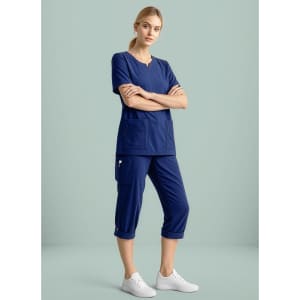 A woman wearing navy scrub top and straight leg scrub pants, paired with white sneakers, standing against a light background.
