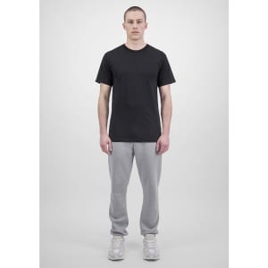 A person wears a black t-shirt and grey track pants, standing against a plain background. The outfit features branding.