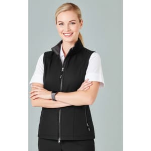A woman in a black vest stands with her arms crossed, smiling against a neutral background. The vest features a zip.