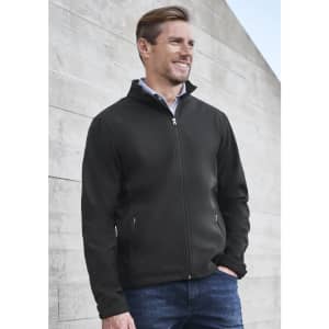 A black soft shell jacket with a zip and side pockets, worn by a man outdoors against a textured wall.
