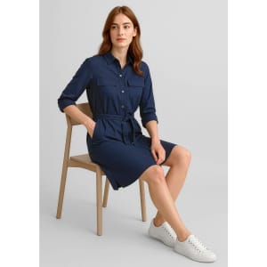 A navy blue dress with two pockets, roll-up sleeves, a belt, and a model seated on a wooden chair.