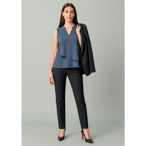 A woman wearing a sleeveless blue top and slim leg black pants, holding a black blazer.