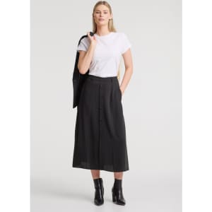 A woman stands wearing a white t-shirt and a black midi skirt, paired with ankle boots. The skirt features buttons and has a logo.
