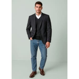 A man in a dark blazer over a black sweater, wearing light blue jeans and brown shoes, stands against a pale background.