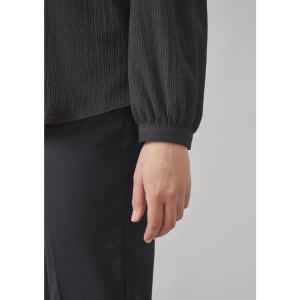 A black long sleeve blouse featuring a ruched texture and gathered cuffs, paired with black pants.