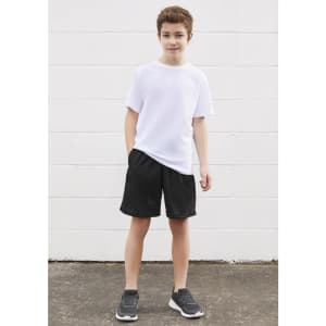 A young boy stands in a white t-shirt and black sports shorts, with hands in pockets, on a concrete surface.