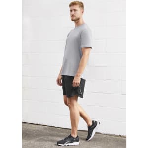 A man wearing a grey t-shirt and black sports shorts, standing against a white wall. The shorts have a logo.