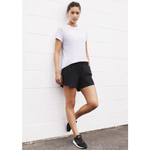 A woman in black sports shorts poses against a white wall, wearing a casual white t-shirt and black footwear.