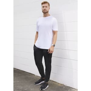 A man wearing a white t-shirt and black track pants stands against a white wall, complemented by black athletic shoes.