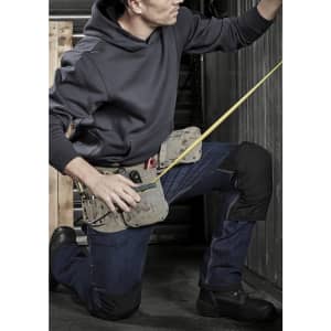 A person kneeling, wearing denim jeans and a dark hoodie, with a tool belt and a measuring tape.