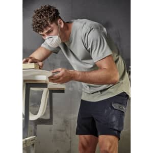 A man in a light green T-shirt and dark shorts, wearing a face mask, works at a table. The shirt has a logo.