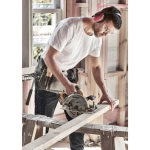A man wearing a white T-shirt uses a power saw on a wooden plank, wearing protective gear and a tool belt.