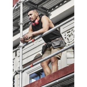 A man wearing a black sleeveless tee, shorts, and a tool belt stands on scaffolding, with headphones around his neck.