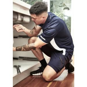 A man in a navy blue and white short sleeve polo is working under a sink, kneeling on a wooden floor.