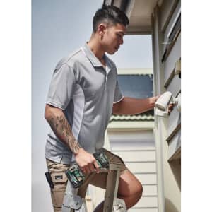 A man in a grey short sleeve polo shirt adjusts a fixture while standing on a ladder, holding tools.