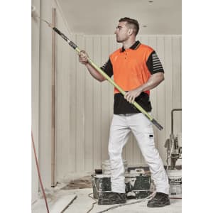 A man in an orange and black short sleeve polo shirt uses a roller to paint a wall. The background has painting equipment.