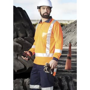 A man wearing an orange long sleeve polo shirt with reflective strips stands on a work site, holding a radio.