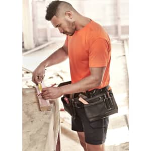 A man wearing an orange hi-vis t-shirt and a tool belt measures wood on a worksite.