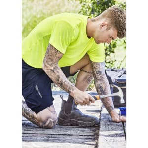 A vibrant yellow men's hi-vis tee worn by a tattooed man, kneeling and using a hammer on a wooden surface.