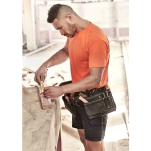 A man in an orange high visibility tee is measuring wood, wearing a tool belt adorned with various tools.