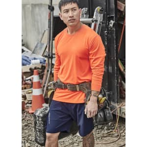 An orange long sleeve safety tee is worn by a man with a tool belt, set against a cluttered worksite background.